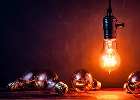 Leuchtende Glühbirne vor einer schwarzen Wand. (iStock.com/ ThomasVogel)